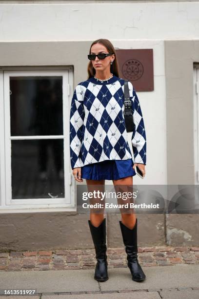 Guest wears black sunglasses, silver earrings, a white and navy blue checkered print pattern wool pullover from Baum und Pferdgarten, a navy blue...