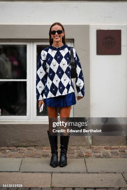 Guest wears black sunglasses, silver earrings, a white and navy blue checkered print pattern wool pullover from Baum und Pferdgarten, a navy blue...