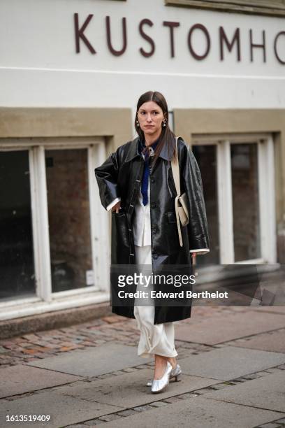 Guest wears silver large earrings, a navy blue shirt, a white shirt, a black shiny varnished leather long coat, a beige matte leather shoulder bag, a...