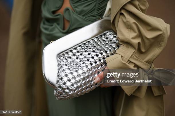 Guest wears a pale khaki long trench coat, a silver shiny braided leather clutch, a green cut-out pattern long dress , outside OperaSport, during the...