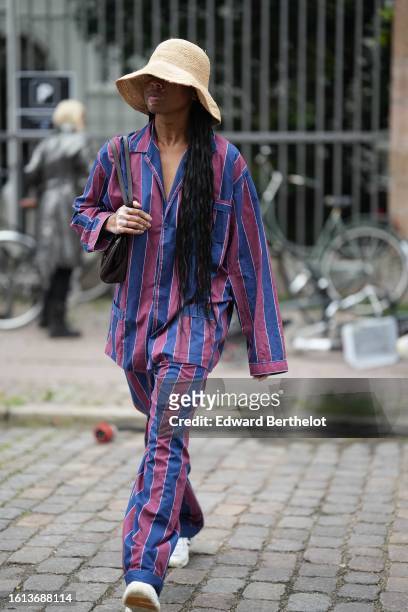Guest wears a beige wickers hat, a navy blue and burgundy striped print pattern oversized cotton shirt, matching navy blue and burgundy striped print...