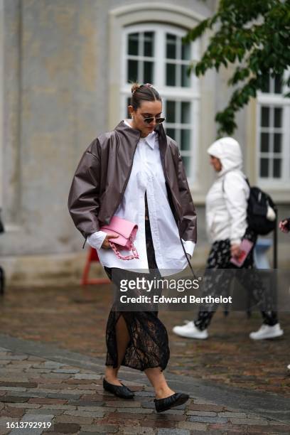 Guest wears black sunglasses, a white oversized shirt, a dark brown shiny leather jacket, a black lace print pattern midi skirt, a pale pink shiny...