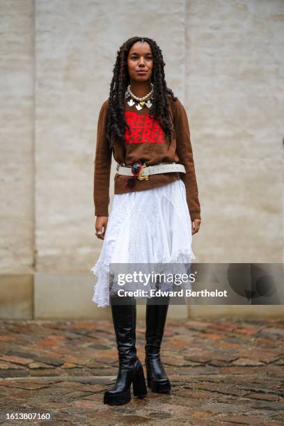 Guest wears a white large pearls with silver pendants necklace, a dark brown with embroidered red pattern pullover, a white latte matte leather belt,...