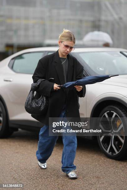 Guest wears brown sunglasses, a dark gray t-shirt, a black oversized blazer jacket, a black shiny leather braided Jodie handbag from Bottega Veneta,...