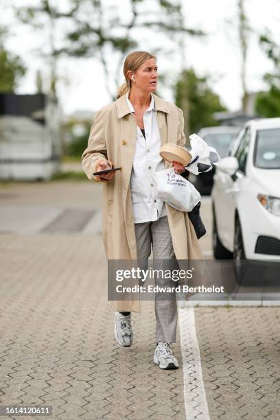 Guest wears a white shirt, a beige long trench coat, a white plastic handbag, silver rings, beige with small black checkered print pattern suit...