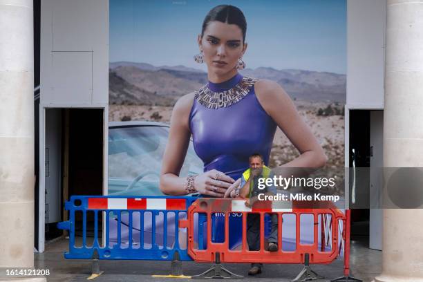Workman at a high end fashion shop refurbishment takes a cigarette break beside barriers in front of a large scale hoarding of a fashion model...