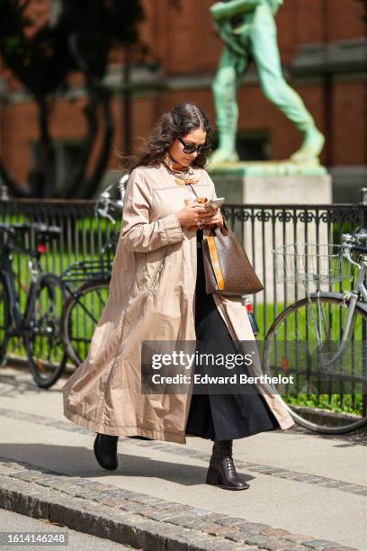 Guest wears black sunglasses, a beige long coat, a dark brown shiny leather large handbag, a black long dress, black socks, black shiny leather block...