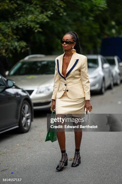 Guest wears black sunglasses, gold large earrings, a pale yellow with black collar blazer jacket, a matching pale yellow shirt skirt, a green shiny...