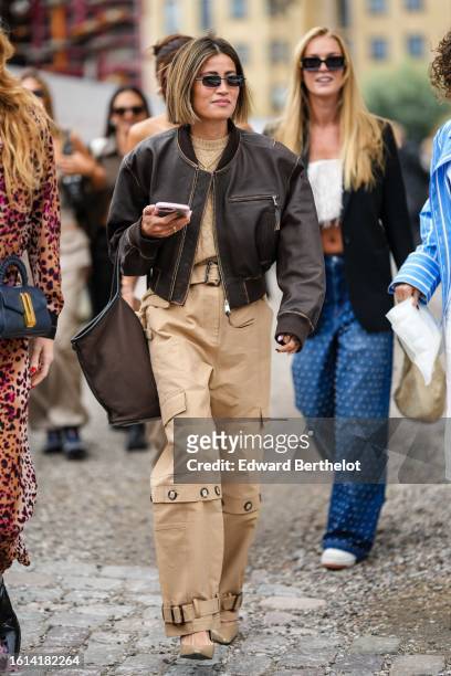 Guest wears black sunglasses, a beige braided wool pullover, a dark brown shiny leather zipper bomber coat, a brown handbag, beige belted cargo...