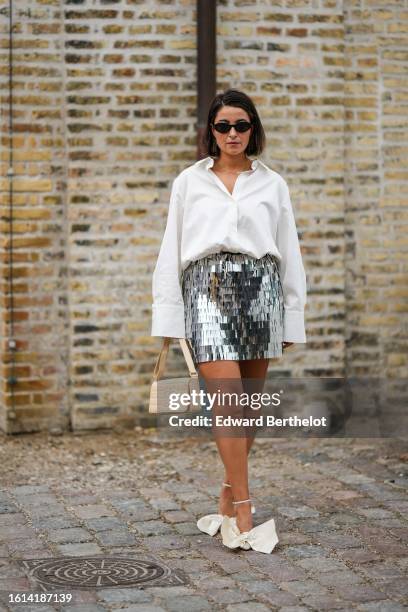 Guest wears black circle sunglasses, a white shirt, a silver mirror fringed short skirt, a beige shiny leather crocodile print pattern vintage...