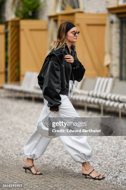 Guest wears a black large headband, black circle sunglasses, a black shiny varnished leather bomber coat, a silver shiny leather crocodile print...