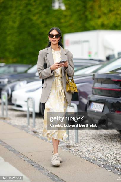 Guest wears black sunglasses, a pale gay long blazer jacket, a pale yellow with white embossed pattern long dress, a yellow shiny leather long...