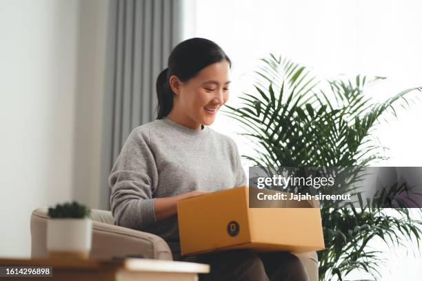 asian woman holding carton box happy excited sitting on armchair in living room at home. - receiving feedback stock pictures, royalty-free photos & images