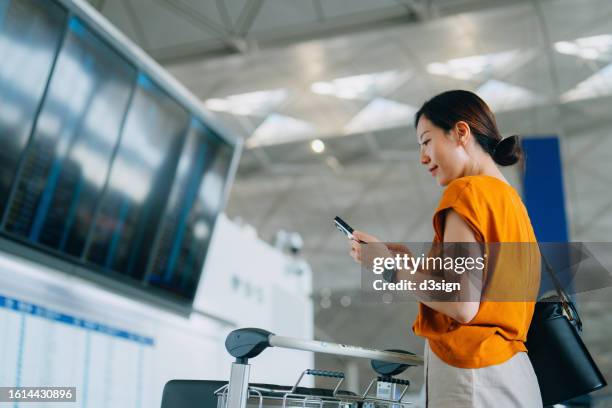 young asian woman checking her flight details on smartphone in front of arrival departure board at airport terminal. checking in with mobile phone. ready for a trip. business travel. travel and vacation concept - departure board front on stockfoto's en -beelden