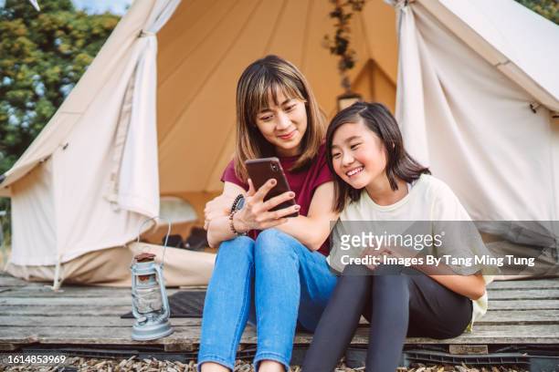 mom & daughter using smart phone together while sitting on the wood deck outside a glamping bell tent - ドームテント ストックフォトと画像