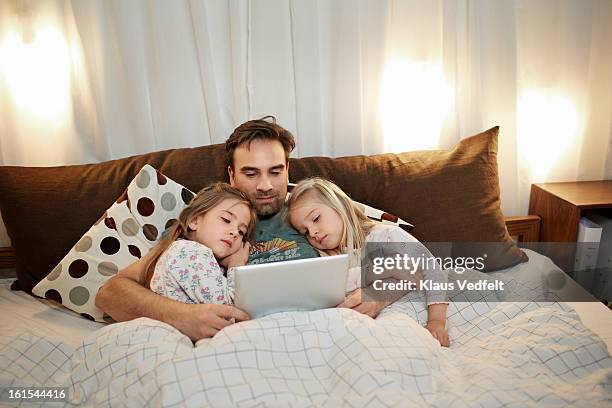 father & 2 girls reading bedtime story on tablet - two men in bed cuddling stock-fotos und bilder