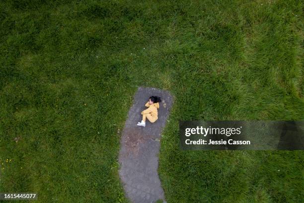 drone shot of girl in yellow tracksuit lying curled up on concrete path edged by grass - shy stock pictures, royalty-free photos & images