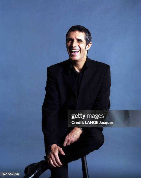 Photo Studio Of Julien Clerc. Julien CLAIR souriant, assis, veste et pantalon noir.