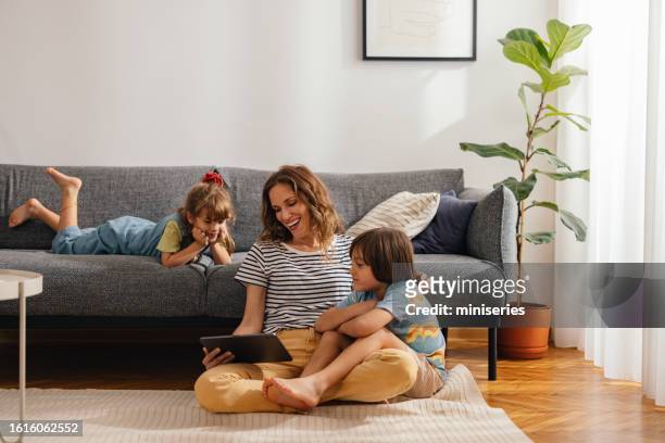moments heureux en famille: la mère est assise par terre et regarde quelque chose sur la tablette numérique avec ses enfants - happy family at home photos et images de collection