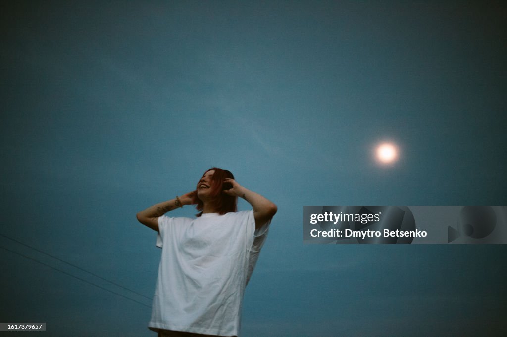 Happy young woman listening music in headphones against the sky at dusk