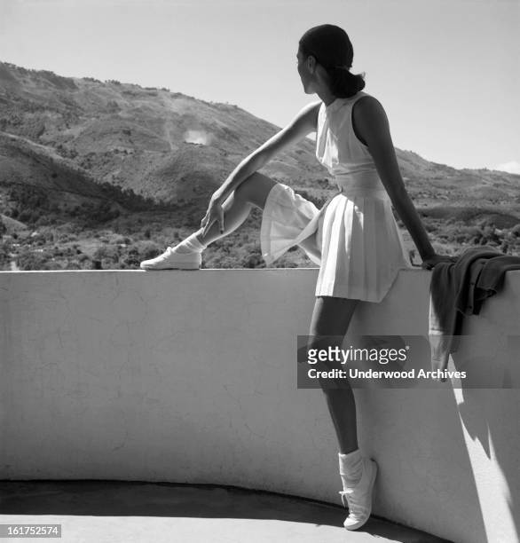 Woman wearing a tennis outfit sitting on a wall, looking at the mountains behind her, February 1947.