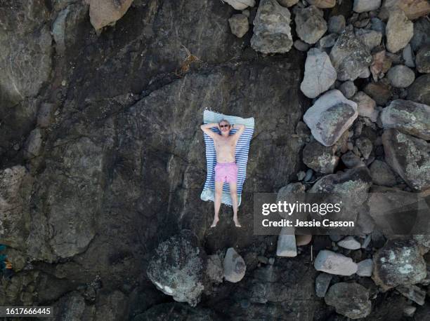 drone shot of mid-life bearded man in swimming shorts lying on back in rocky ground - midlife crisis stock pictures, royalty-free photos & images