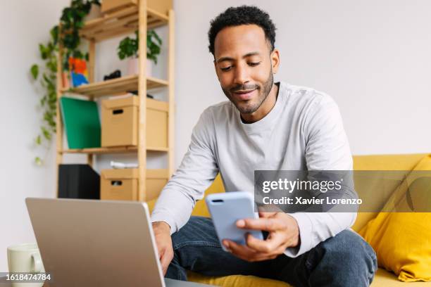 happy young african american smiling guy looking at cellphone screen while using laptop computer sitting on yellow sofa at comfortable living room. - conto corrente foto e immagini stock