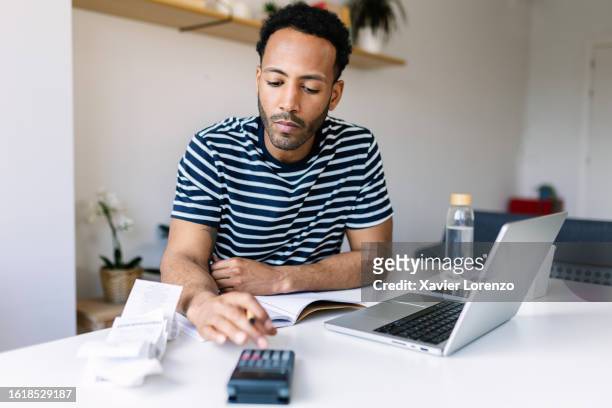 young adult man with laptop checking bills, taxes, bank account balance and calculating expenses sitting at living room table. - calculadora fotografías e imágenes de stock