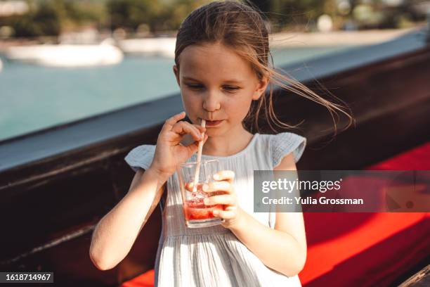 a cute girl in a blue dress drinks delicious juice on the street from a glass glass through a straw. - niño-tomando-agua fotografías e imágenes de stock