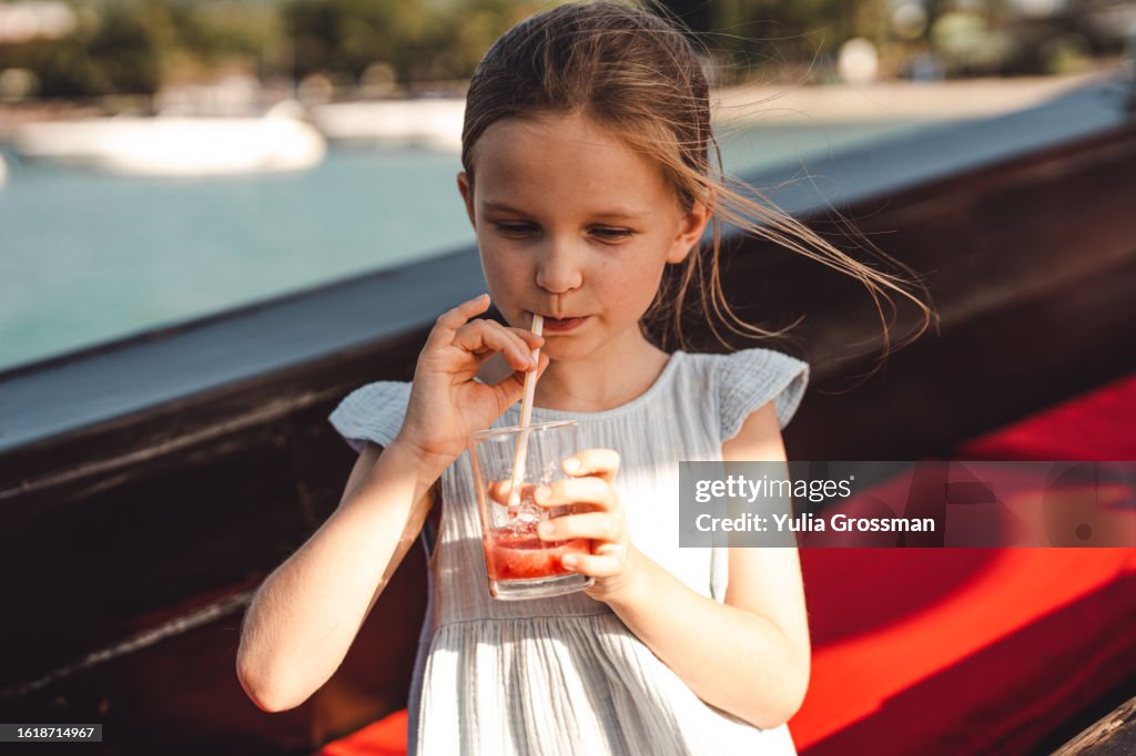 A cute girl in a blue dress drinks delicious juice on the street from a glass glass through a straw.
