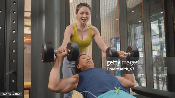 a 65-year asian muscular athlete man  in a blue outfit using a long pull machine for his strength training, focusing on the chest and back muscles in a gym, building up his body muscle happily with support from an asian mid-adult woman fitness instructor - 39 year old stockfoto's en -beelden