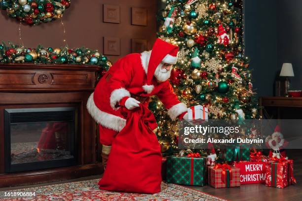 santa claus with big bags of gifts puts a gift under the christmas tree. christmas concept - santa claus fotografías e imágenes de stock