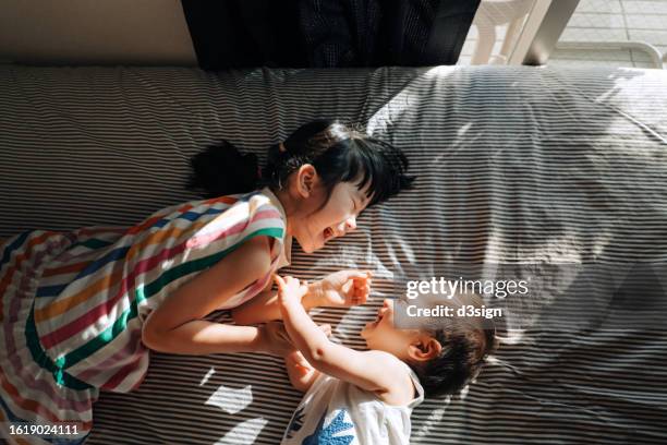 directly above shot of happy little asian girl and her baby sister having fun together and playing on the bed, both of them are smiling joyfully. siblings love. love, bonding and togetherness - japanese tickle - fotografias e filmes do acervo