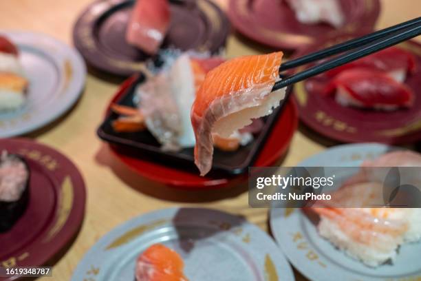 Posed photograph of a pair of chopsticks holding up a piece of sushi next to plates of sushi inside a Japanese restaurant on August 23, 2023 in Hong...