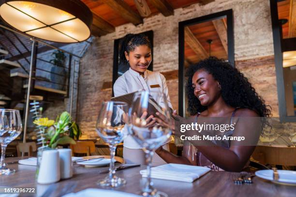 afro-haired latina woman waitress serves an afro-latin woman who goes to the restaurant to try delicious dishes - caribbean dream stock pictures, royalty-free photos & images