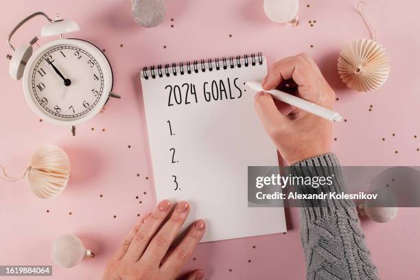 woman's hand writing goals 2024 for new year. new year routine. pink background with festive decoration. flat lay style - new year's day stockfoto's en -beelden