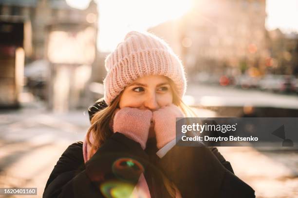young woman warming her hands while standing on the street in city during cold weather in winter - warme kleidung stock-fotos und bilder