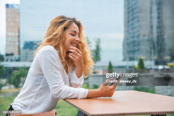 frau mit lockigem haar sitzt in einem café und schaut mit einem lächeln auf das telefon - office smile light stock-fotos und bilder