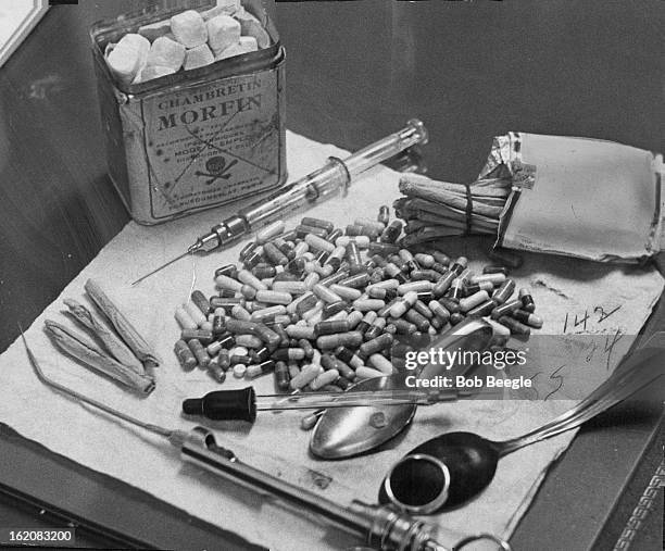 Confiscated narcotics and implements used by addicts are shown at Morals Bureau headquarters. Can contains a morphine compound. Capsules hold heroin....