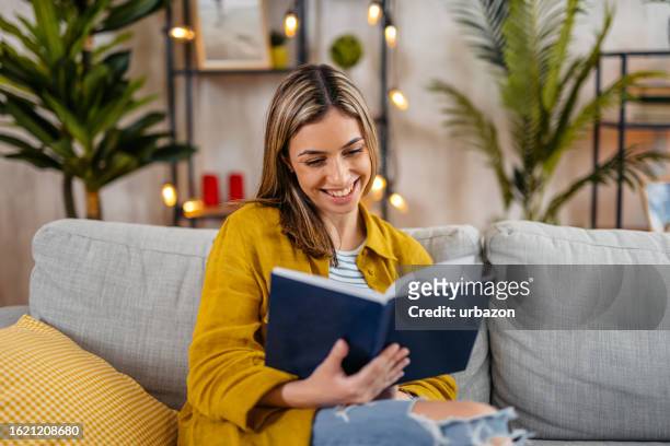 donna che si siede sul divano e legge un libro a casa - sfogliare libro foto e immagini stock