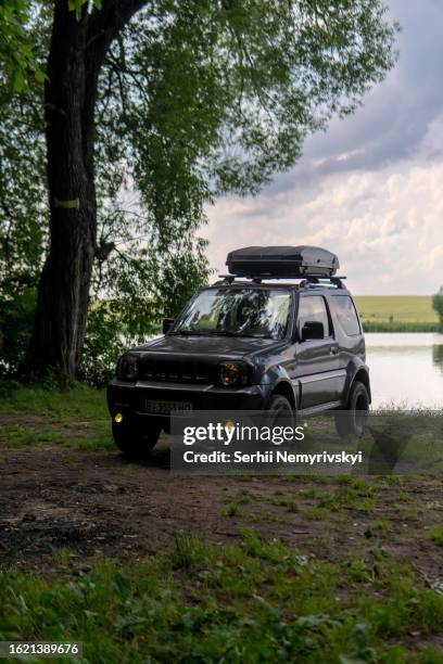 khmelnytskyi, ukraine - july 2, 2023: old suzuki jimny 2010 jb43 with black roof rack box on the top, outdoor, traveling by car. rainy summer day on the shore of the lake, forest. vertical photo - car roof box stock-fotos und bilder