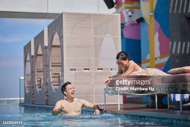 asian man splashing water playfully at attractive asian female in hotel pool - the weekend in news around the world stock pictures, royalty-free photos & images