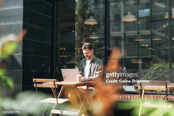 brunette young man in casual clothes using mobile phone working on laptop sitting in cafe at the city - café bar fotografías e imágenes de stock