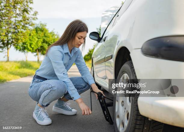 una mujer joven dedicada al mantenimiento de neumáticos con un gato de neumáticos de automóvil en la carretera - rueda pinchada fotografías e imágenes de stock