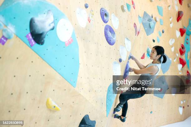 rock and walls climbing teaches you to overcome challenges and body strength. side view of skilled female japanese climber gripping hand and foot holds as she follows the route up a horizontal climbing wall in a gym. - mental toughness stock pictures, royalty-free photos & images