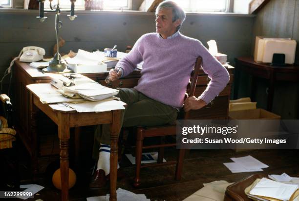 In the paper-strewn writing studio at his summer home, American writer and journalist George Plimpton works at his desk, Wainscott, New York, January...