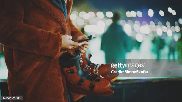 frau in winterjacke hält schlittschuhe in der hand, während sie smartphone an beleuchteter eisbahn benutzt - amsterdam noel stock-fotos und bilder