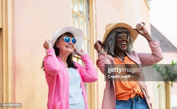 zwei multirassische ältere frauen mit sonnenbrillen und sonnenhüten - happy sprong two persons sun stock-fotos und bilder