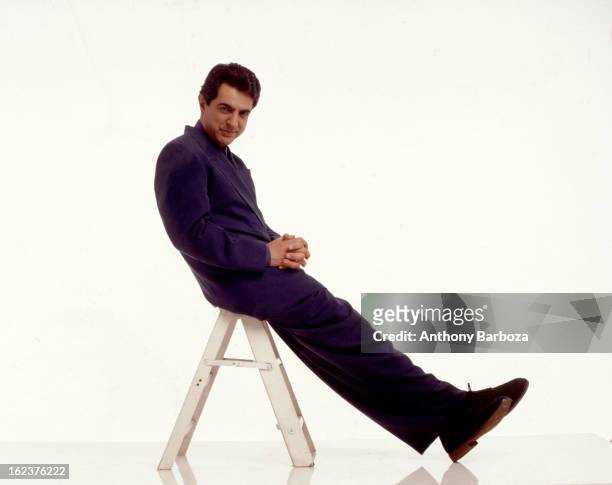 Portrait of American actor Joe Mantegna as he sits on a short step ladder, against a white background, 1991.