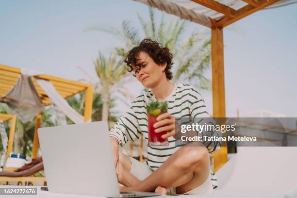 online learning and cocktail party on the beach: short haired woman enjoys cocktail and work on laptop for e-learning while basking in the sun on a tropical beach - online happy hour stock pictures, royalty-free photos & images